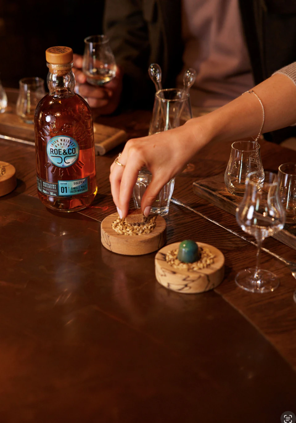 A hand reaching for a cork coaster near a bottle of Roe & Co whiskey on a wooden table with glasses and whiskey paraphernalia.