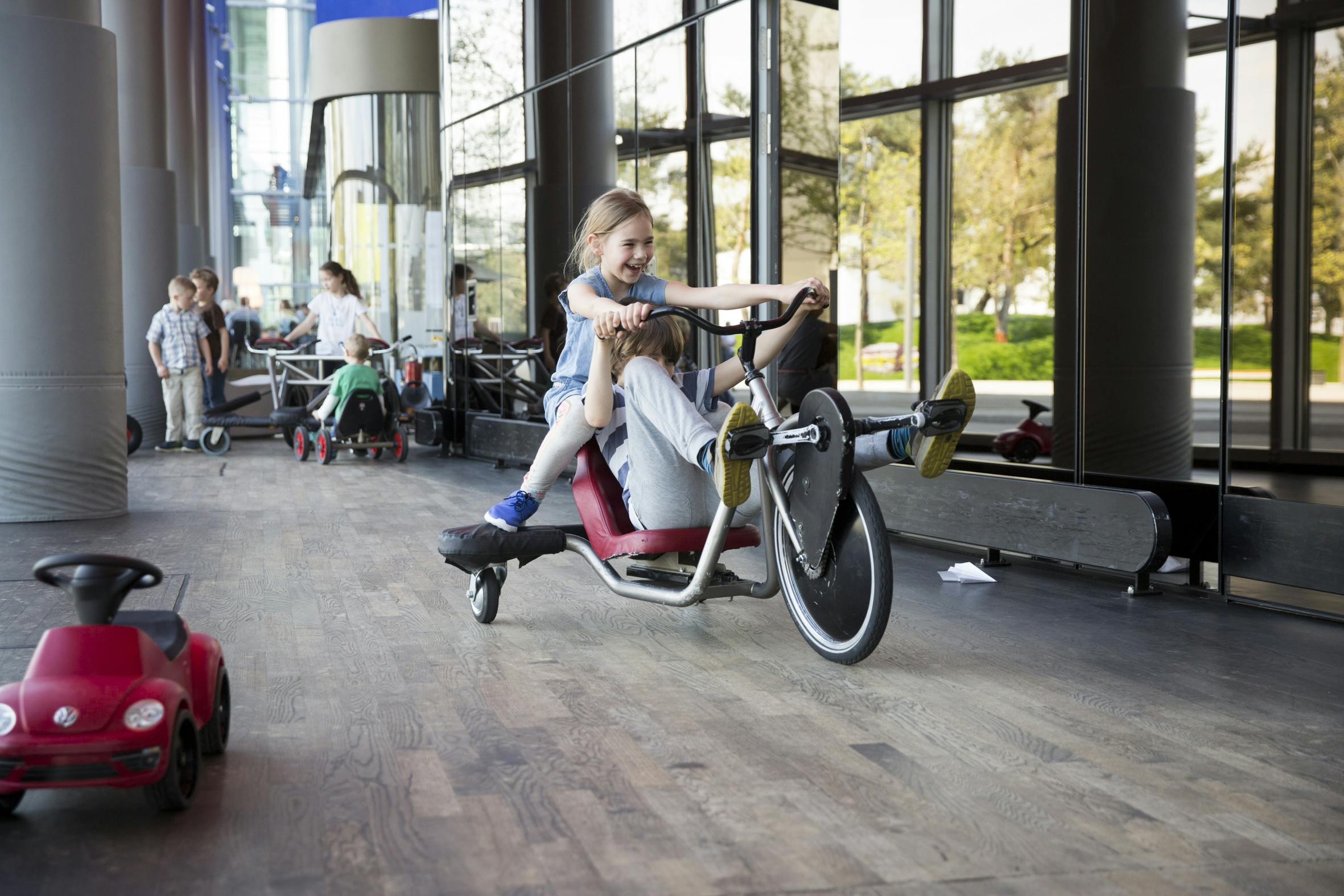 Two children riding a three-wheeled bike indoors. Other children in the background, and large windows revealing trees outside.