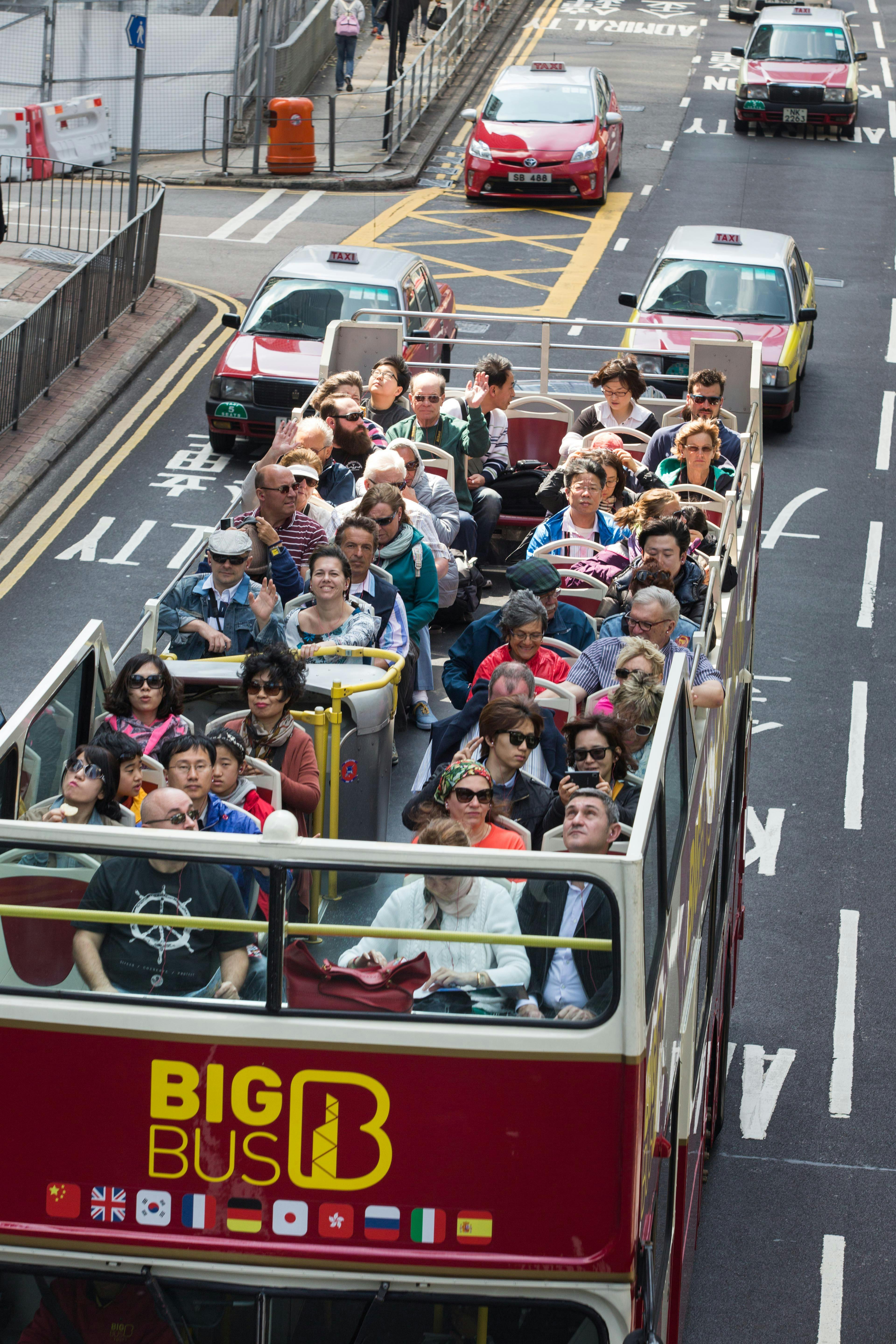 上階のデッキに座っている乗客で満たされた2階建てのツアーバスで、タクシーが前方にある市街地を走行しています。