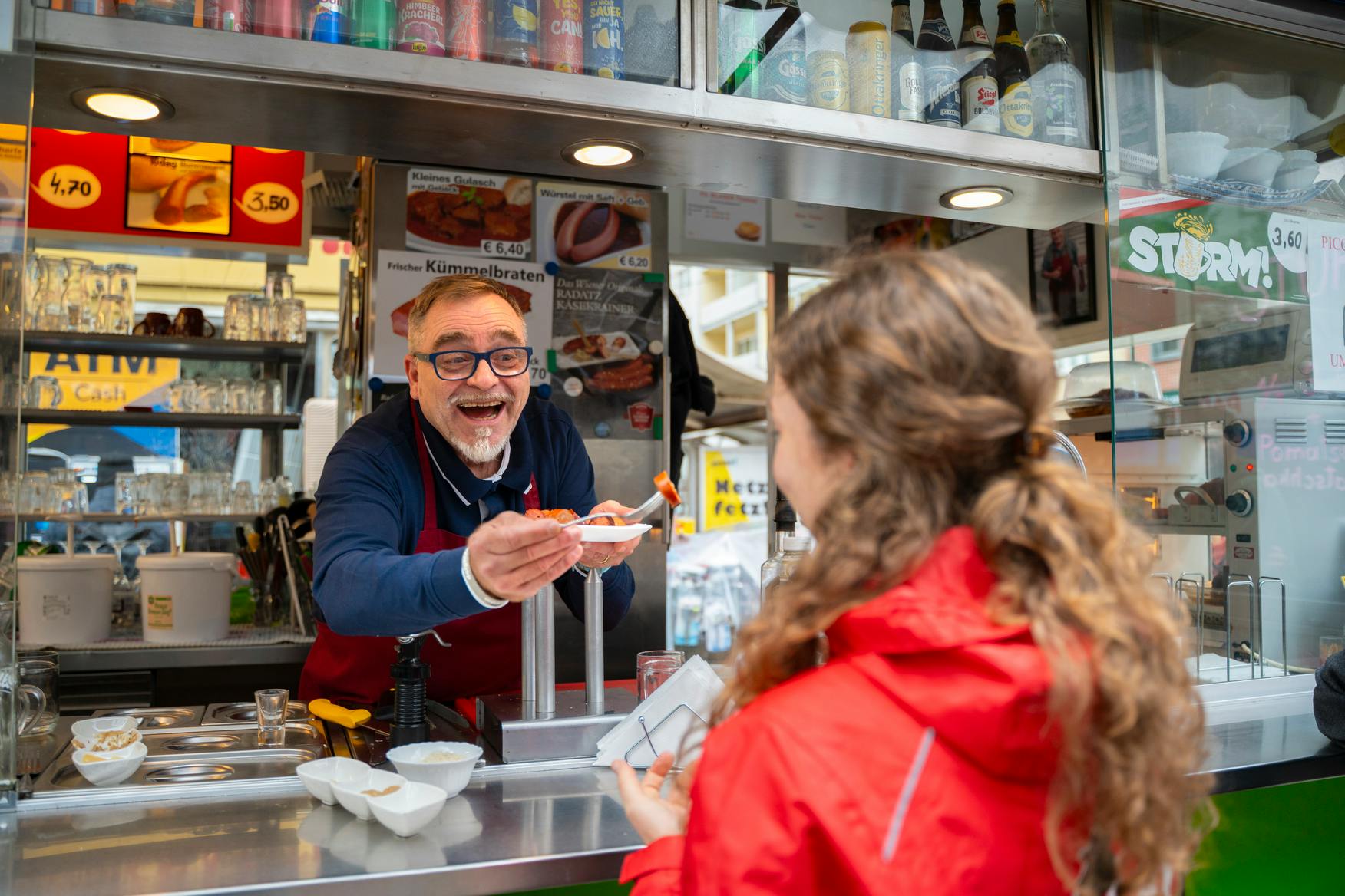Um vendedor sorridente numa banca de comida serve um prato a um cliente de casaco vermelho. Vários itens do menu são apresentados acima.