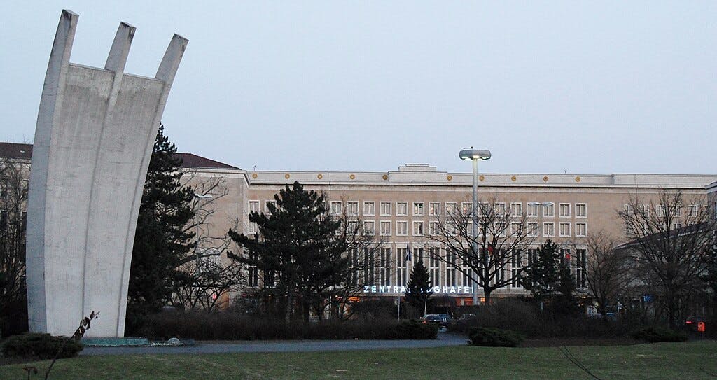 A large building with the words "Zentralflughafen" is seen behind trees and a curved concrete monument in a park.