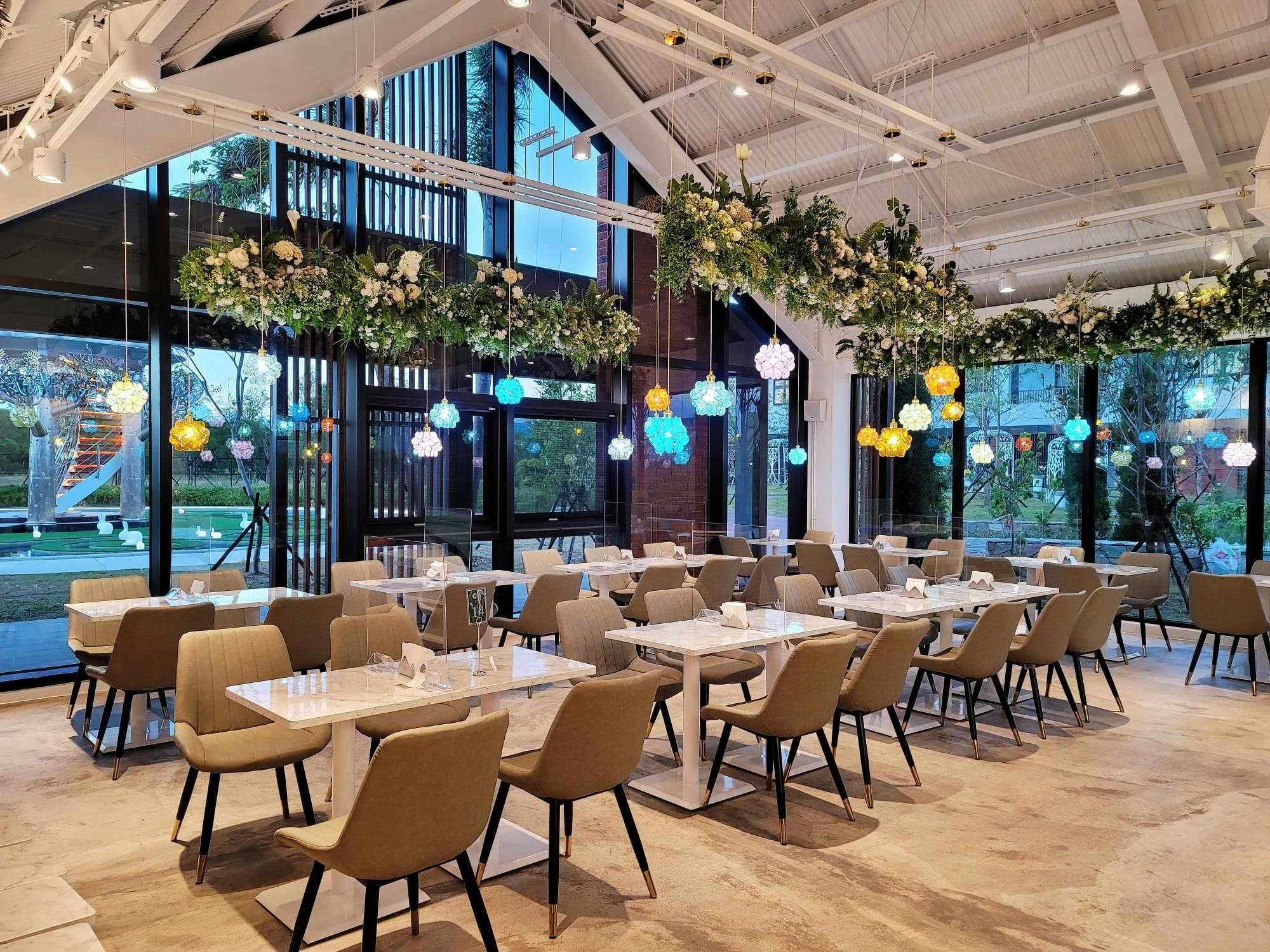 A modern dining area with tables, chairs, colorful hanging lights, and floral arrangements, set in a bright, spacious venue.