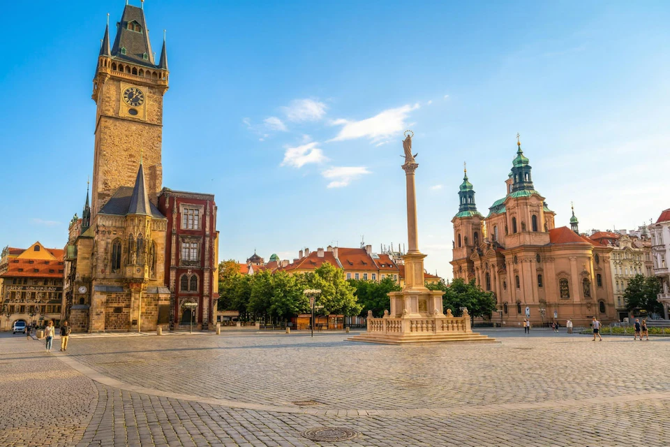 A city square with historical buildings, a tall column with a statue, and a clock tower under a clear blue sky.