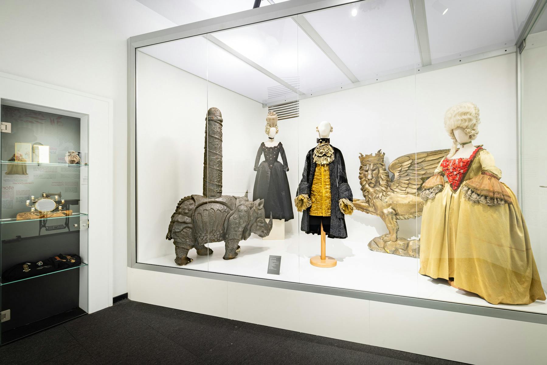 Una mostra museale che espone costumi d'epoca e sculture, tra cui una figura di rinoceronte e un leone alato.