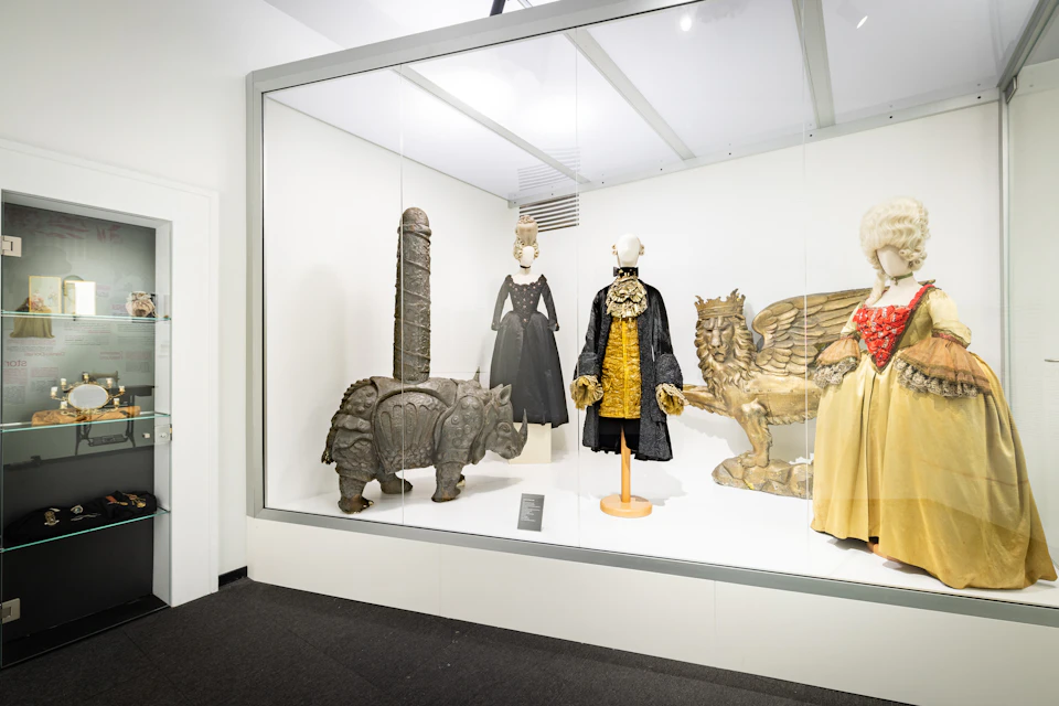 A museum exhibit displaying period costumes and sculptures, including a rhinoceros figure and a lion with wings.