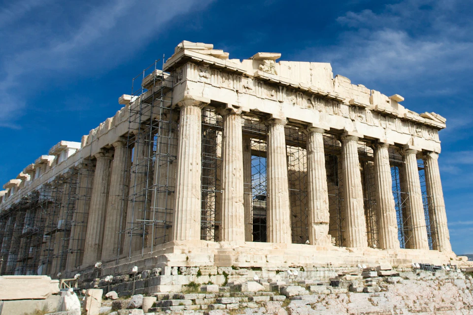 Ancient temple with tall columns and scaffolding on one side under a blue sky. Stone steps lead up to the structure.