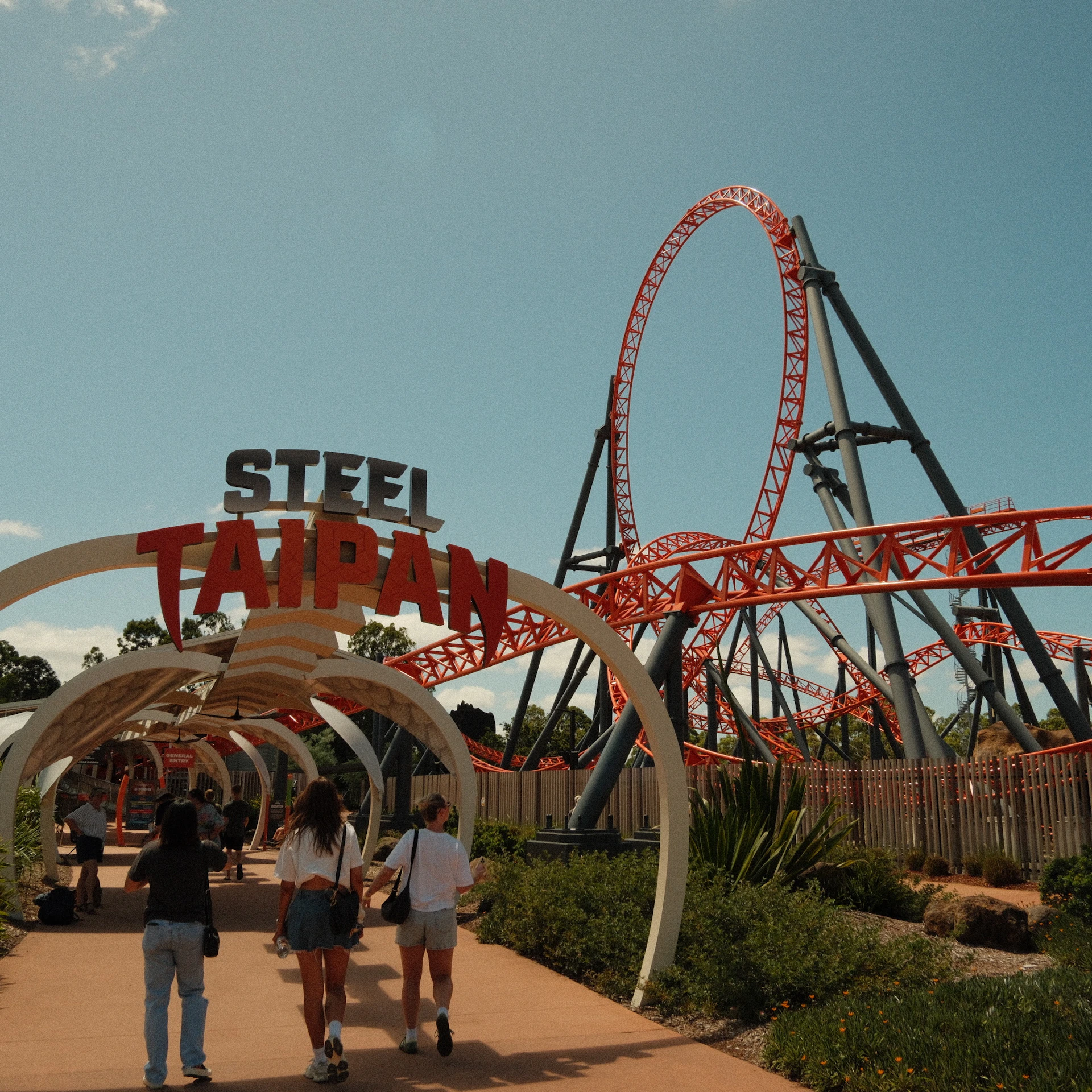 People walking under an archway labeled 'Steel Taipan' towards a red roller coaster on a sunny day.