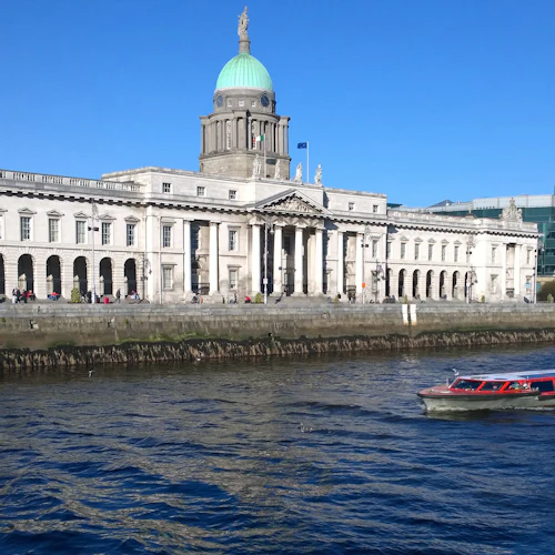 Grå neoklassisk byggnad med en grön kupol vid en flodstrand, en röd och vit båt på det blå vattnet och moderna glasbyggnader i bakgrunden.