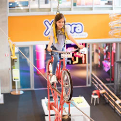 En tjej rider en röd cykel på en upphängd kabel på ett inomhusnöje eller vetenskapscenter, med en säkerhetssele.