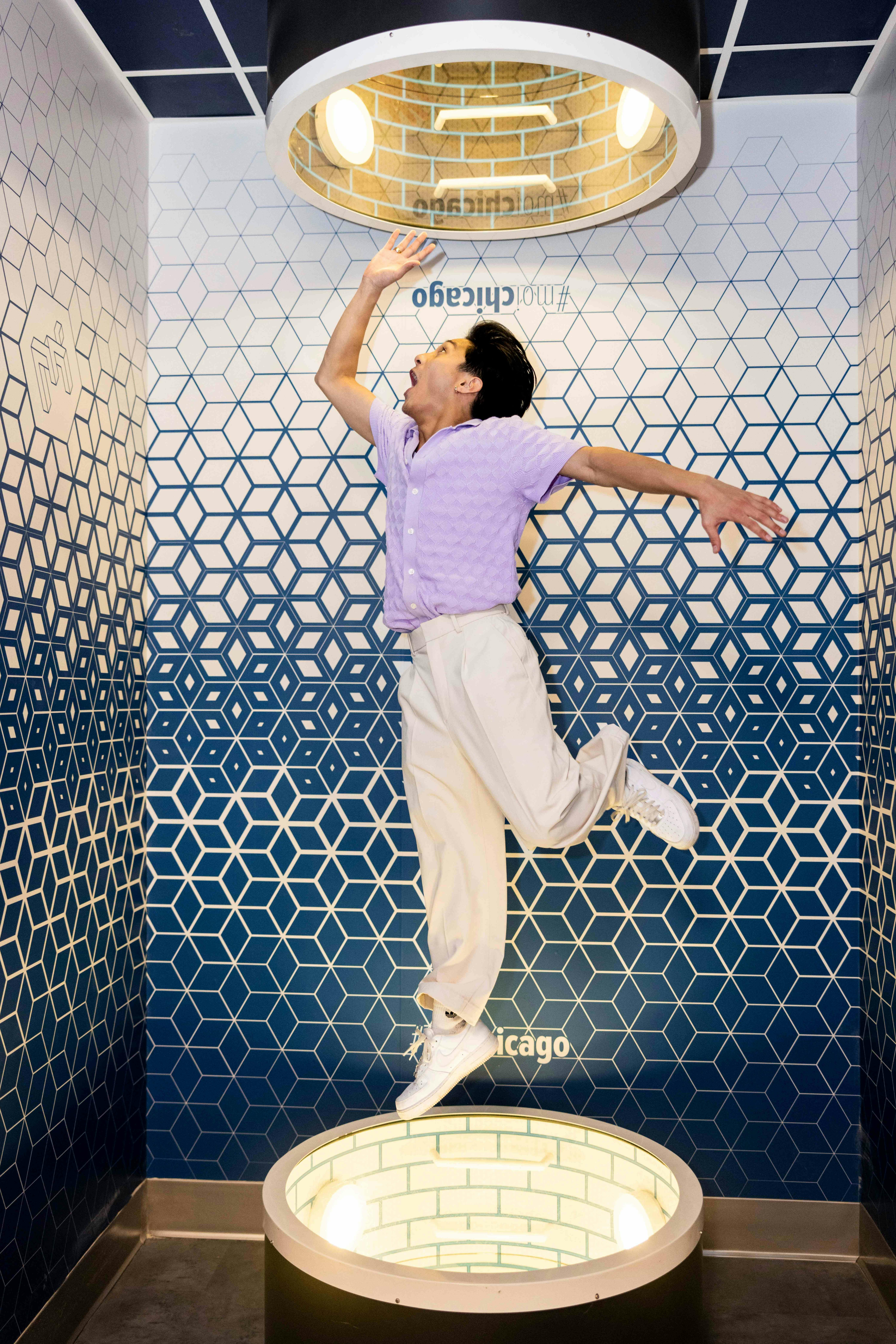 A person in mid-jump against a blue and white geometric hexagon-patterned wall, wearing a purple shirt and white pants.