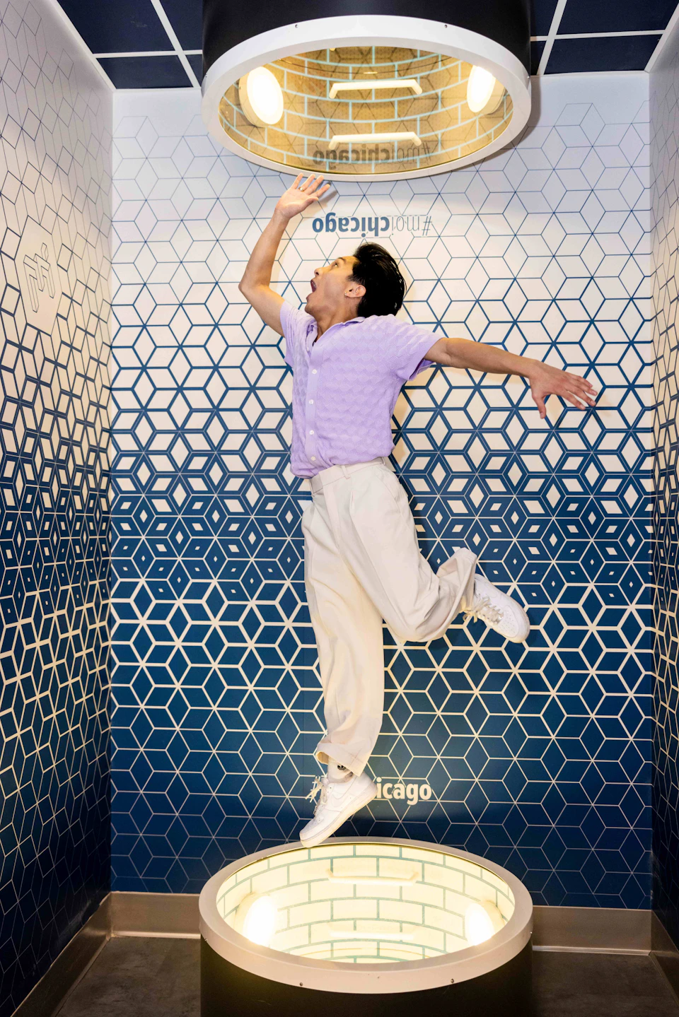 A person in mid-jump against a blue and white geometric hexagon-patterned wall, wearing a purple shirt and white pants.