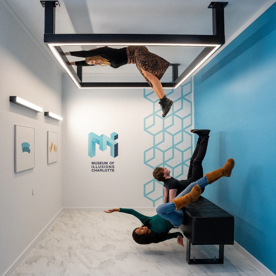 Three people in an upside-down room exhibit, one appearing to walk on the ceiling, at the Museum of Illusions, Charlotte.