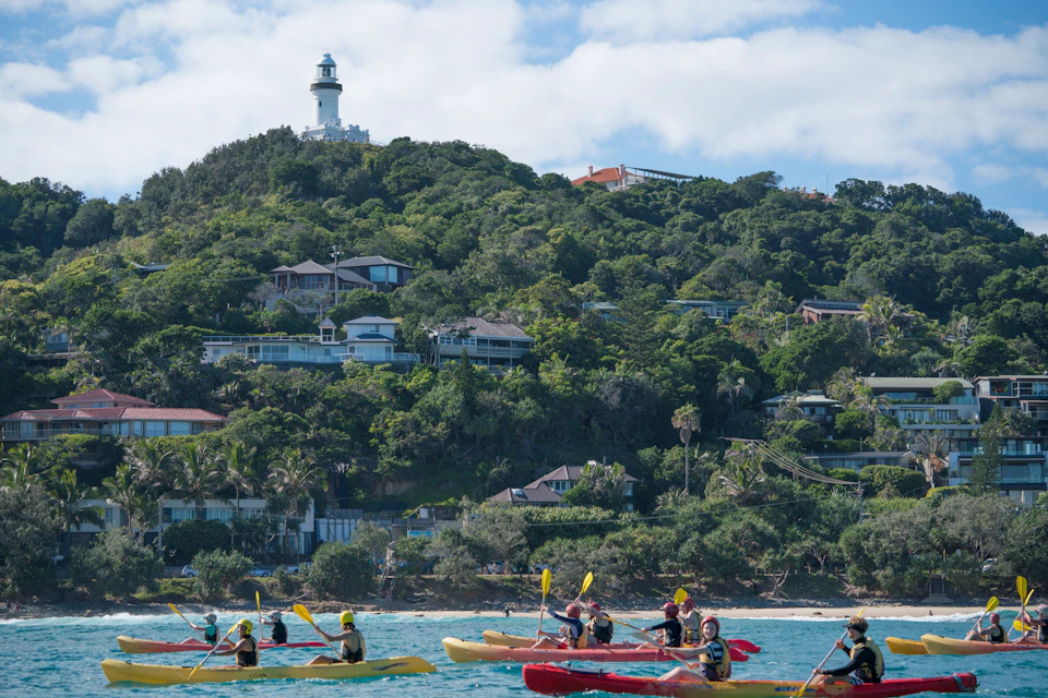 Kayakers paddle near the shore with a forested hill and a lighthouse in the background.