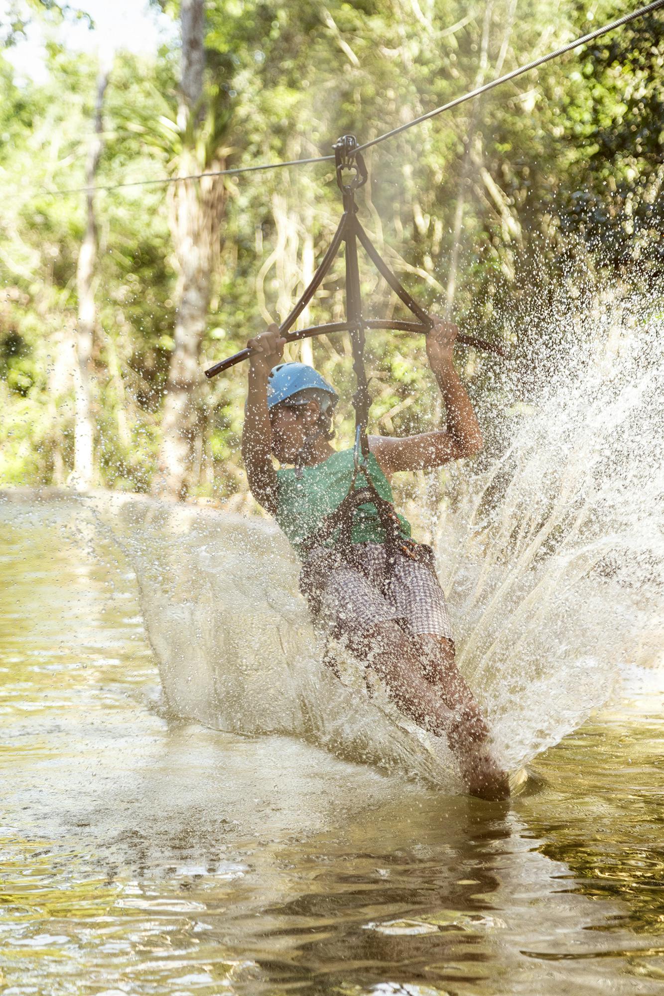 青いヘルメットと緑色のシャツを着た人物が水の上にジップライニングを施し、木を背景に大きなスプラッシュを生み出しています。