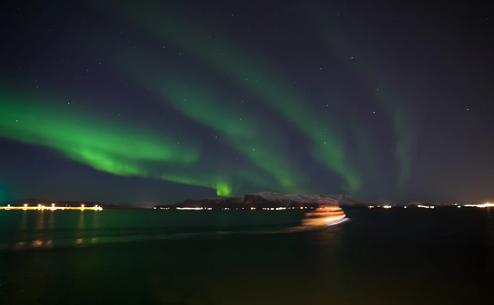 The image shows green Northern Lights over a dark sky and a blurred boat on water with mountains in the background.