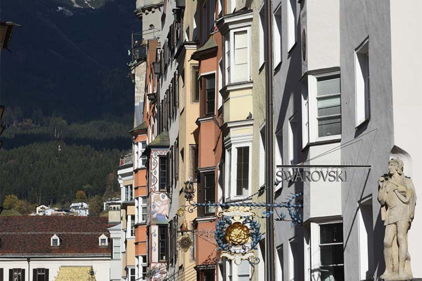 街道上有五颜六色的狭窄建筑，施华洛世奇商店的标志，华丽的悬挂标志，右边还有一个男人的雕像。