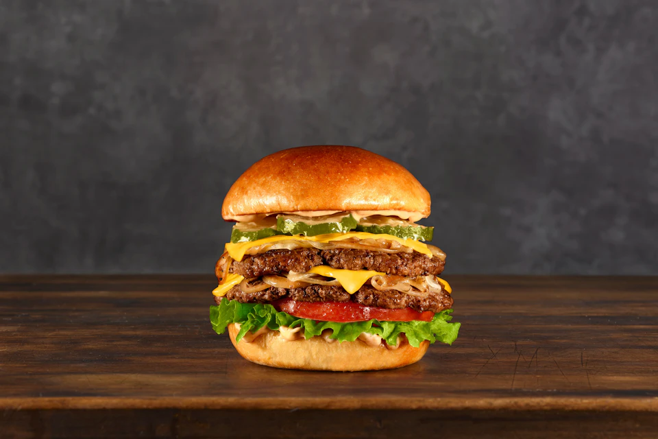 A double cheeseburger with lettuce, tomato, pickles, onions, and melted cheese on a wooden surface against a dark background.