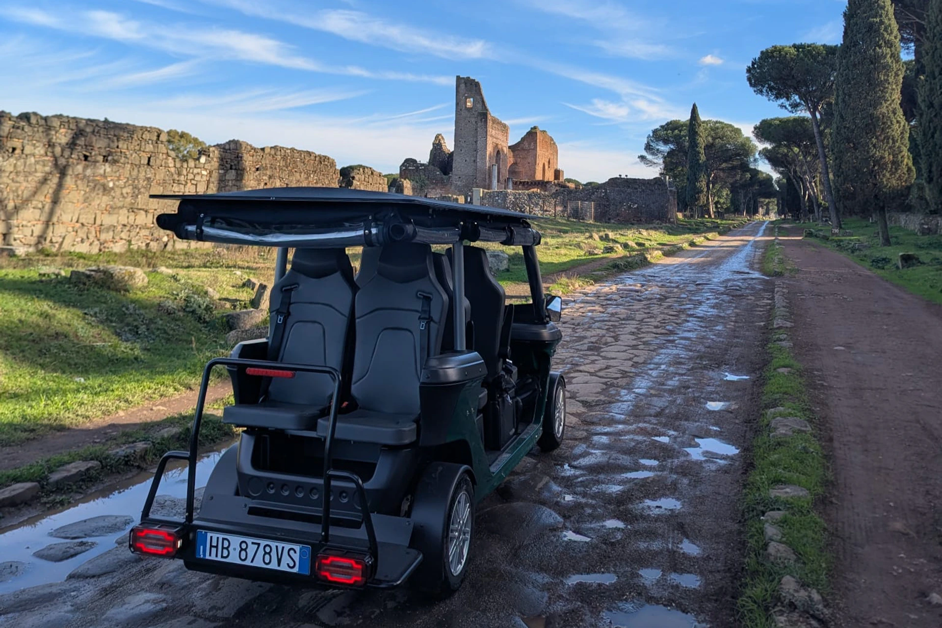 A small green electric vehicle parked on a cobblestone path in a historical area with ruins and greenery under a blue sky.
