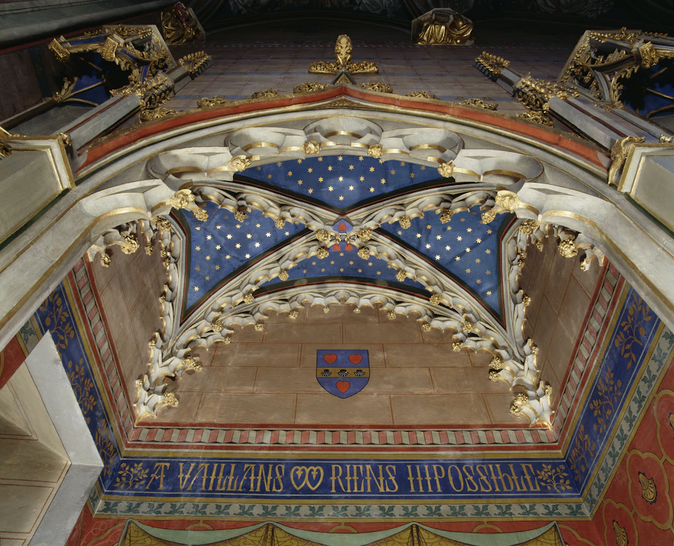 An ornate Gothic ceiling with gold accents, a blue starry sky panel, a crest with fleur-de-lis, and Latin inscription below.