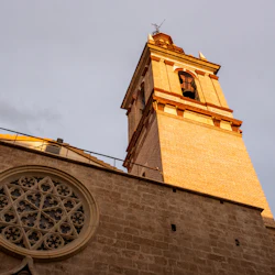 Tegelstenstorn med välvda fönster och ett runt, dekorativt glasmålningsfönster, upplyst av varmt solljus mot en molnig himmel.