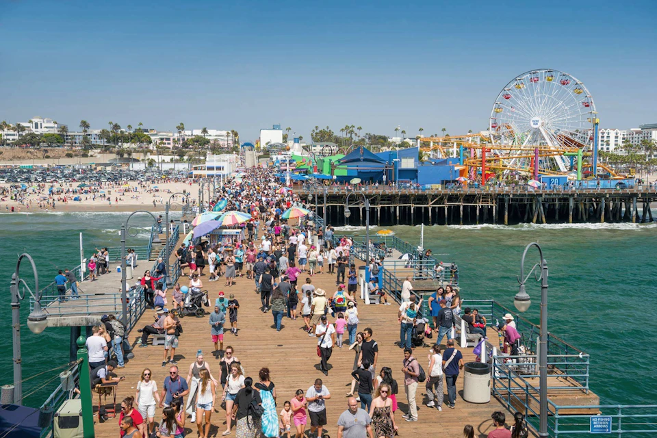 Crowds of people walking on a bustling pier, with a beach, colorful structures, and a Ferris wheel in the background.