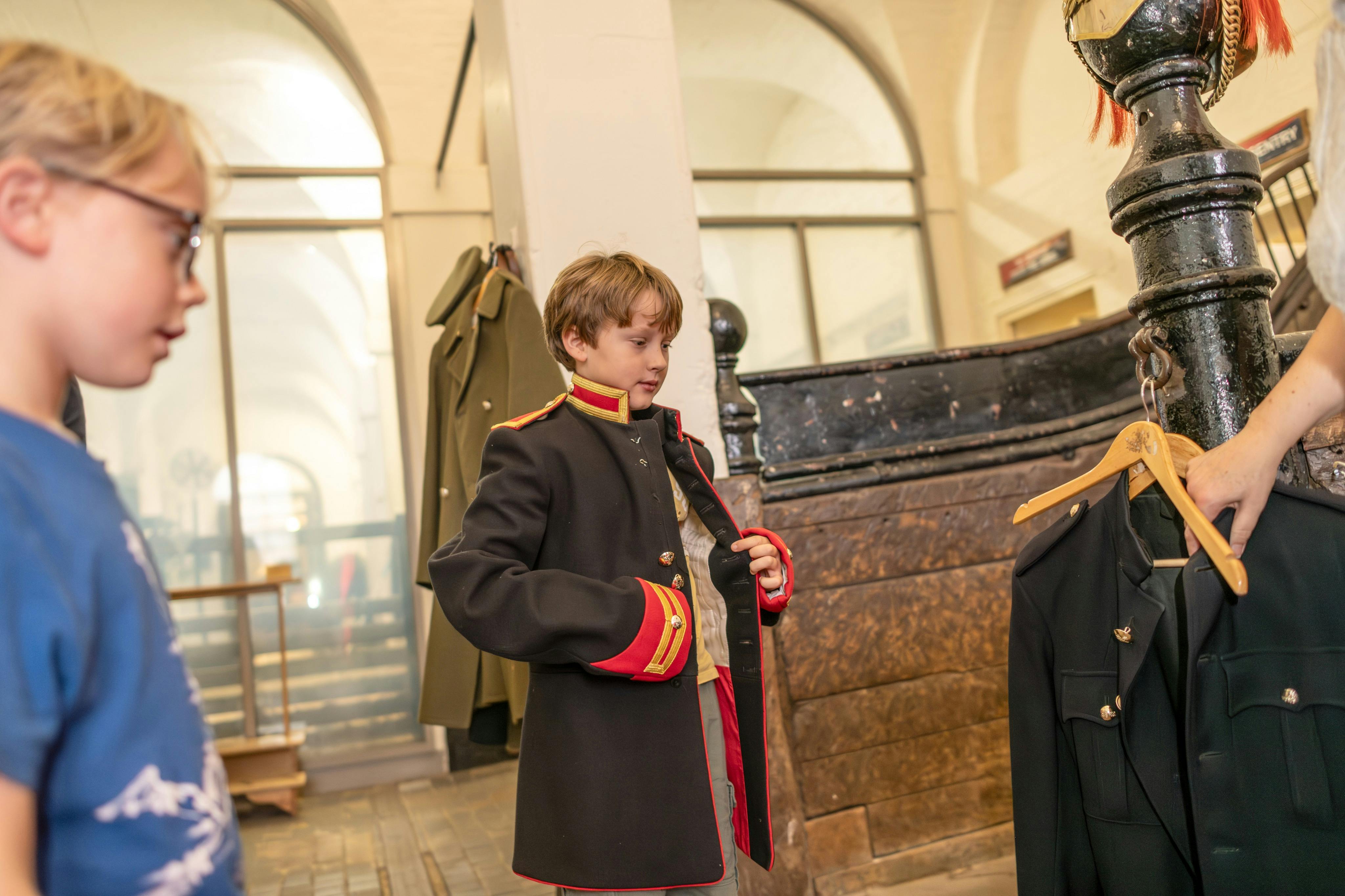 Ett barn försöker sig på en historisk militärstil kappa inomhus medan ett annat barn observerar i en museiliknande miljö.