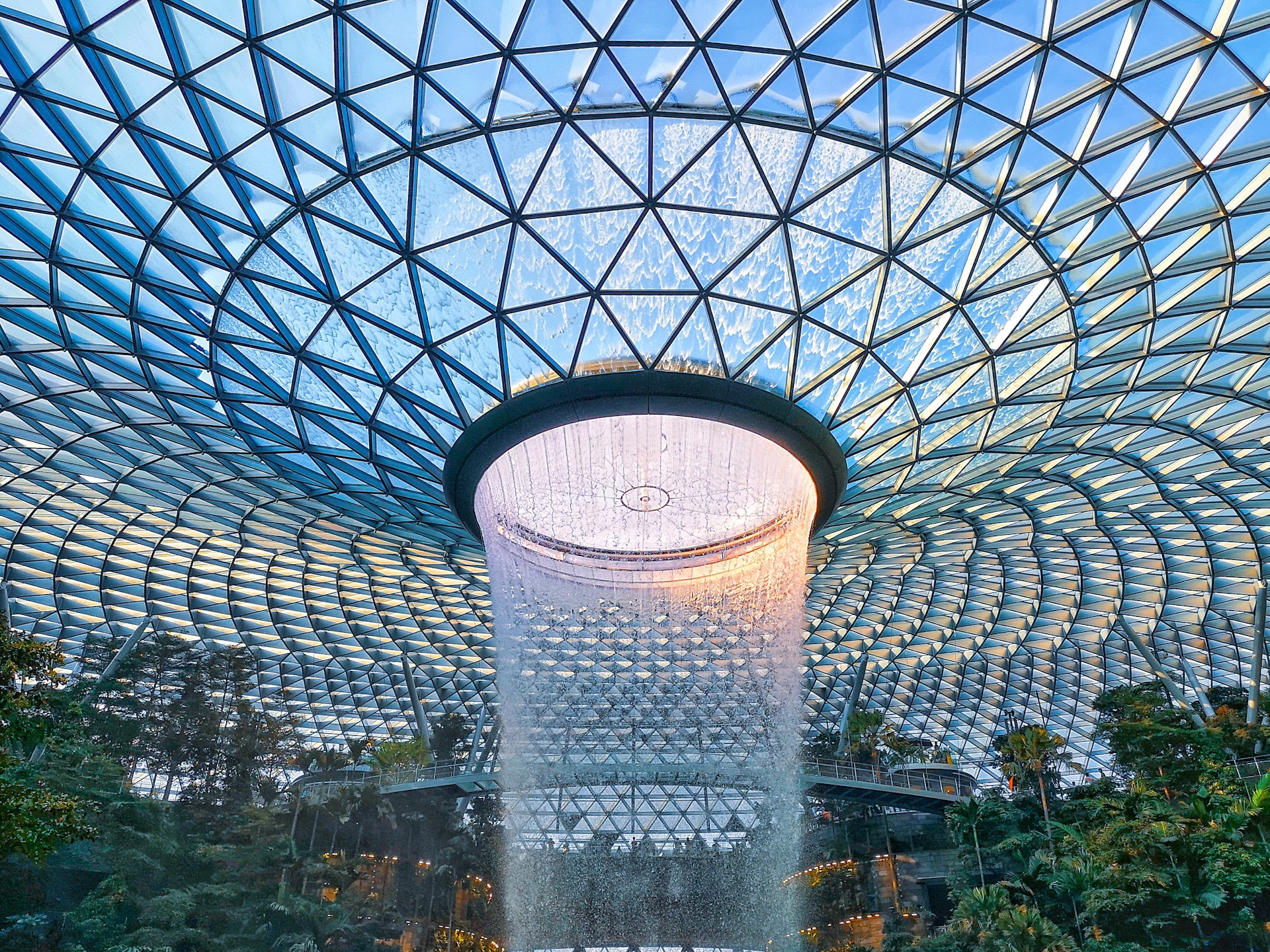 Indoor waterfall under a large, geometric glass dome, surrounded by lush greenery and buildings.