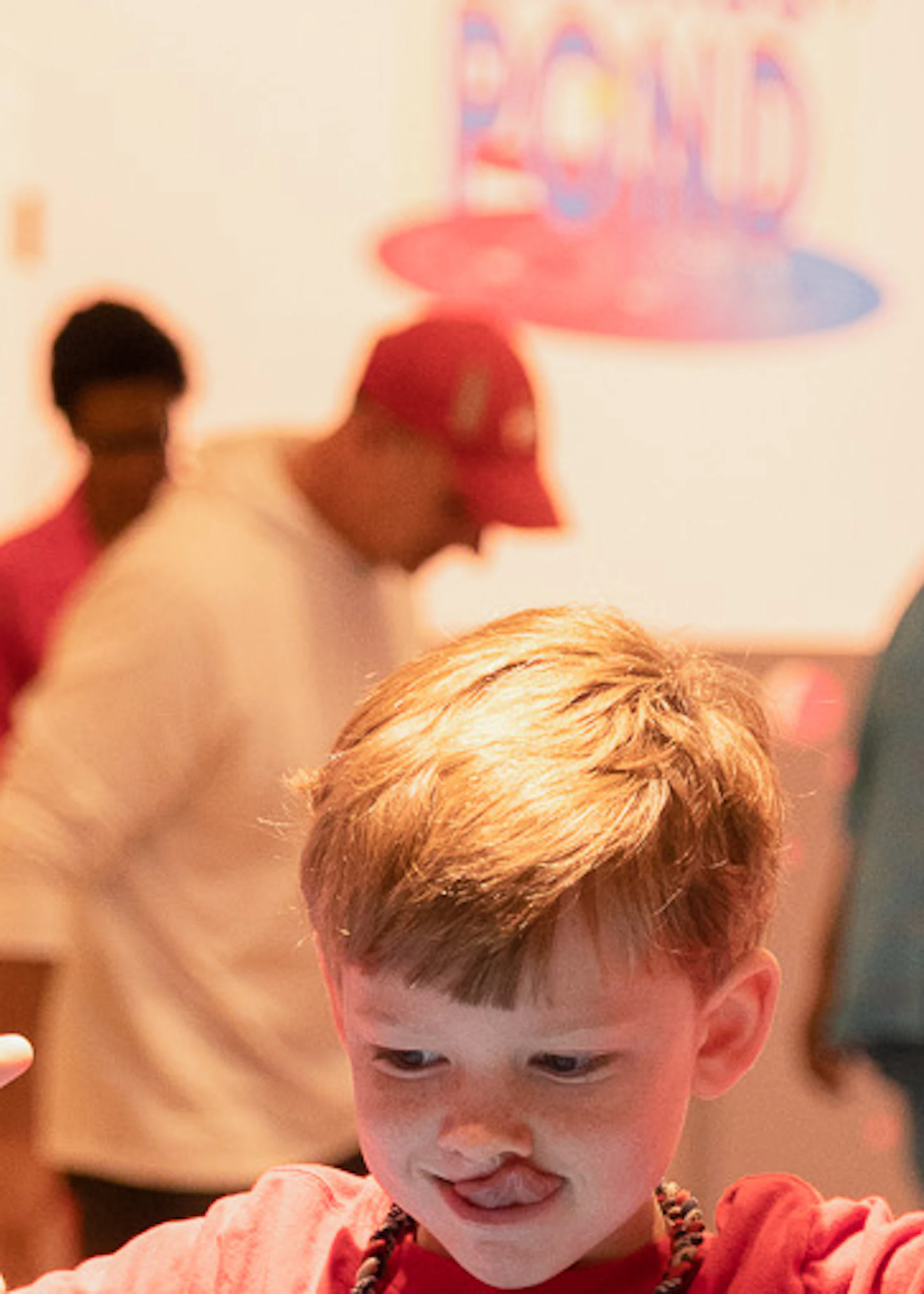 A woman assists a child walking on a blue surface in a decorated room, with other adults and children in the background.
