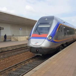 Um comboio moderno prateado com riscas azuis e laranja na plataforma de uma estação, sob um céu nublado. Um indivíduo está de pé à esquerda.