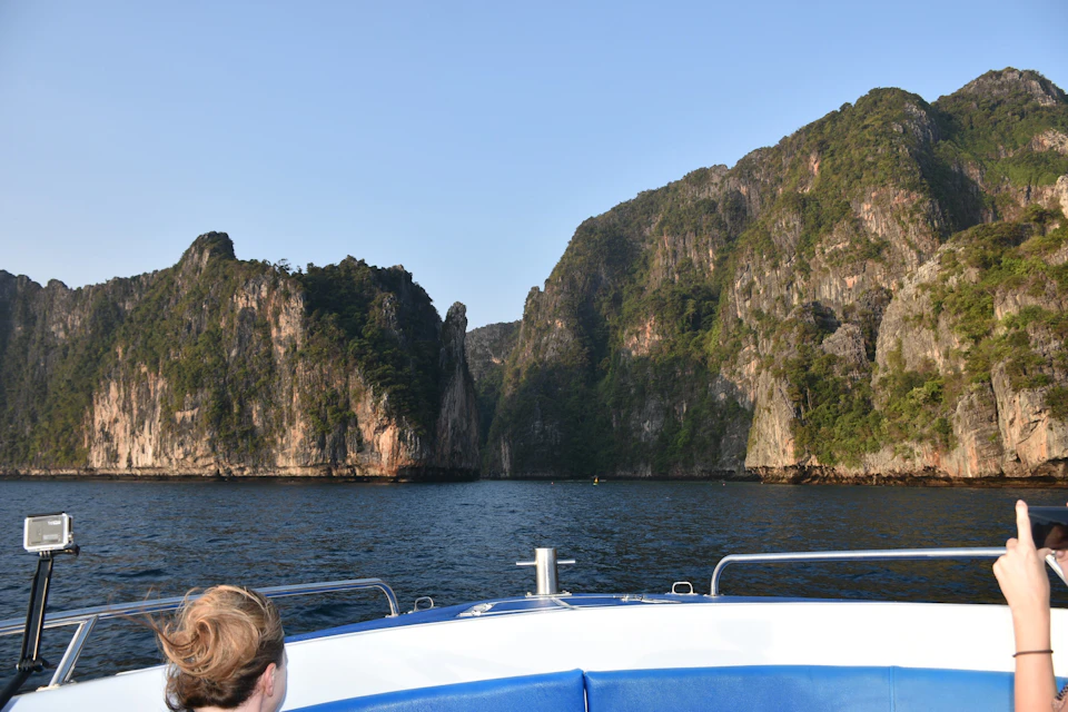 Person on a boat approaching tall, rocky cliffs covered in greenery under a clear blue sky.
