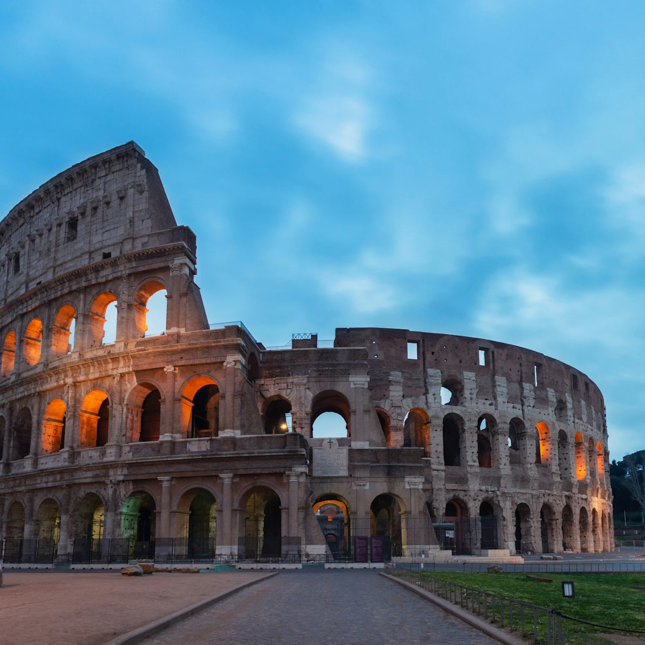 The Colosseum under a blue sky with lights illuminating the arched openings, and an empty pathway leading to the structure.