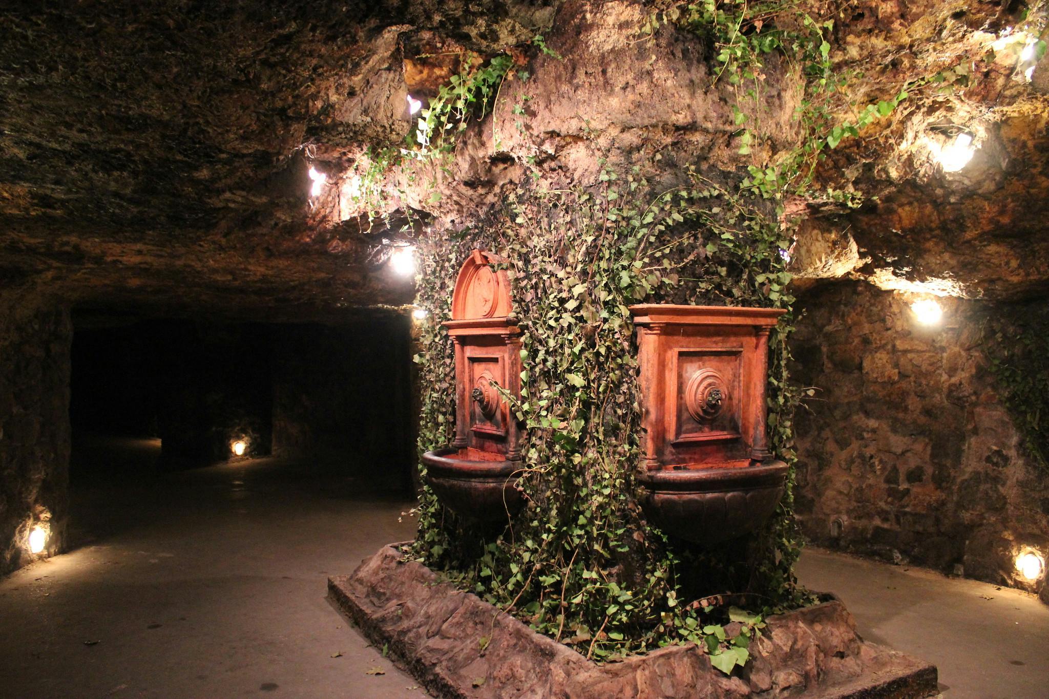 Underground chamber with ivy-covered stone walls, illuminated by small lights, featuring two ornate fountains.