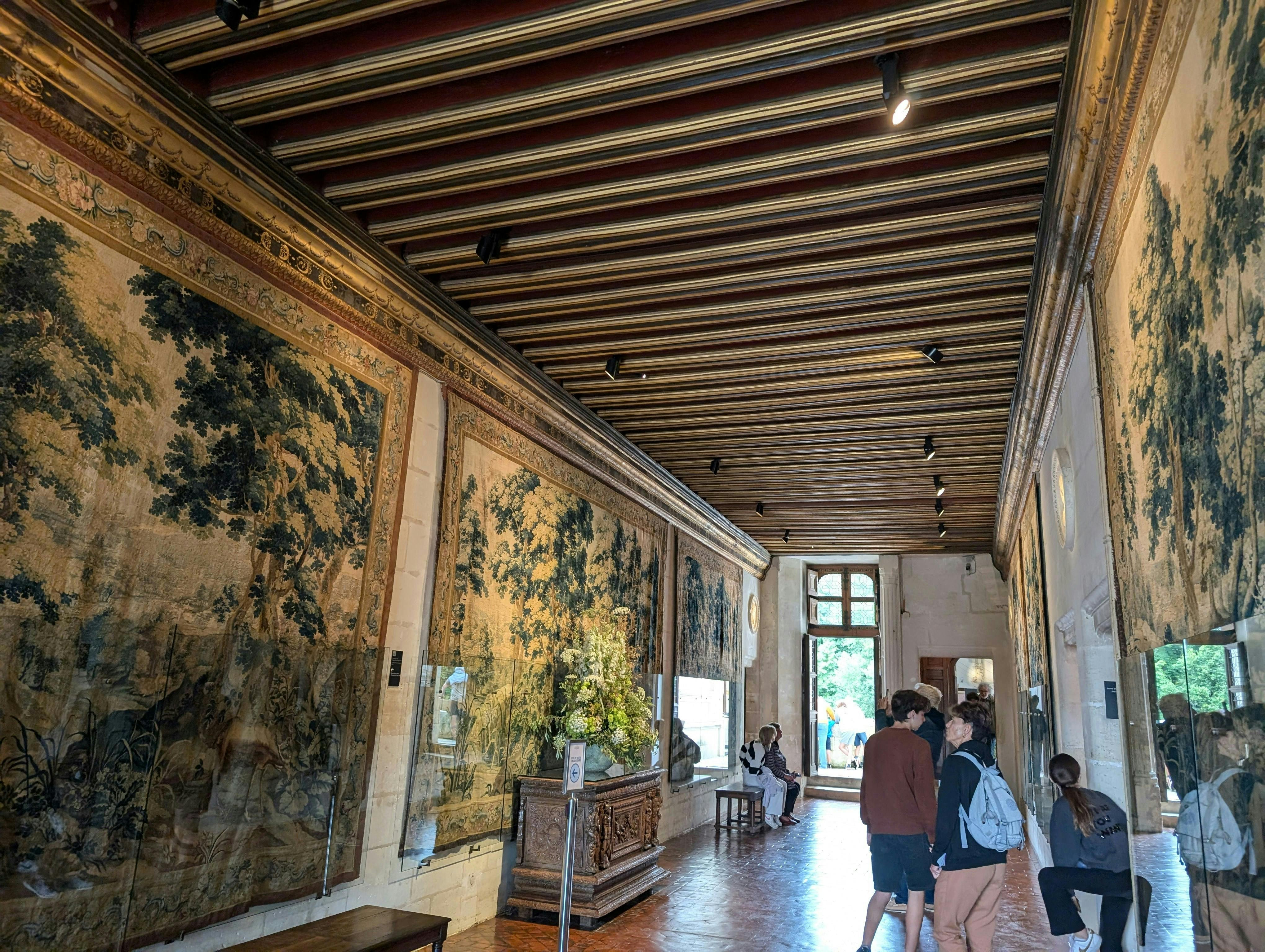 벽에 화려한 태피스트리가 놓인 박물관 복도, 나무 천장, 전시회를 관람하는 몇 명의 사람들.