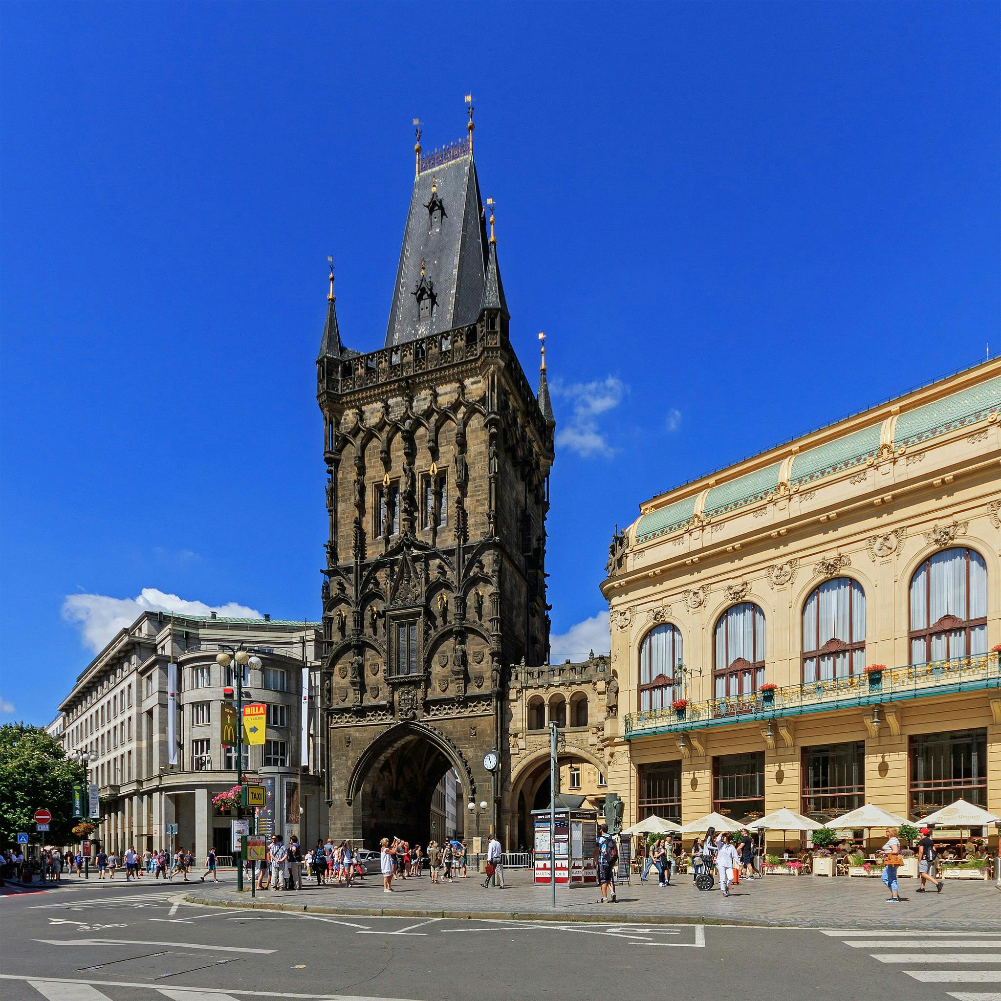 Historical tower with ornate architecture next to a beige building. People walking and sitting at outdoor cafes under a bright blue sky.