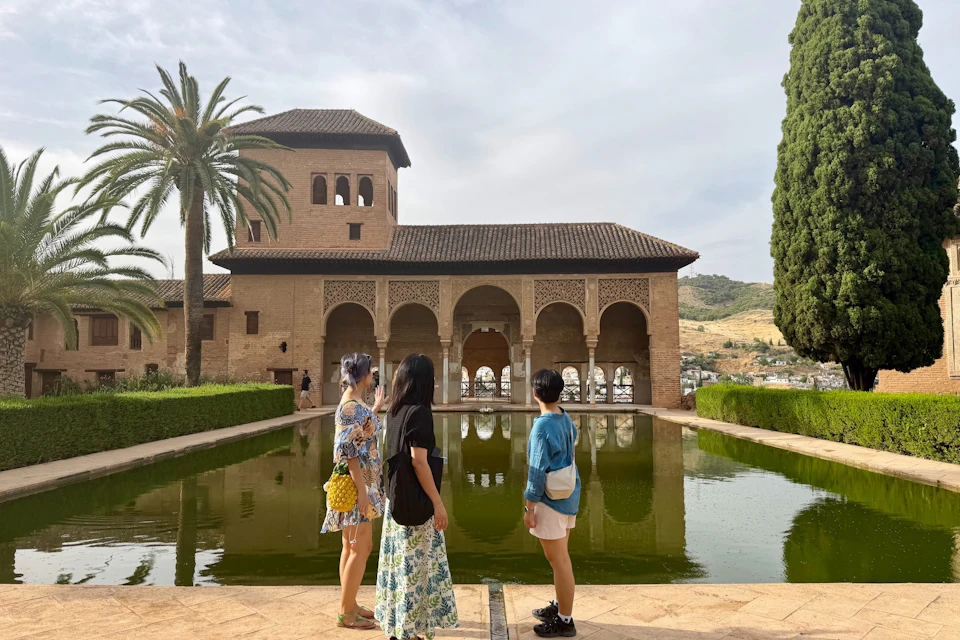 Three people stand near a reflective pool, facing a historic building with arches and intricate designs, surrounded by greenery.