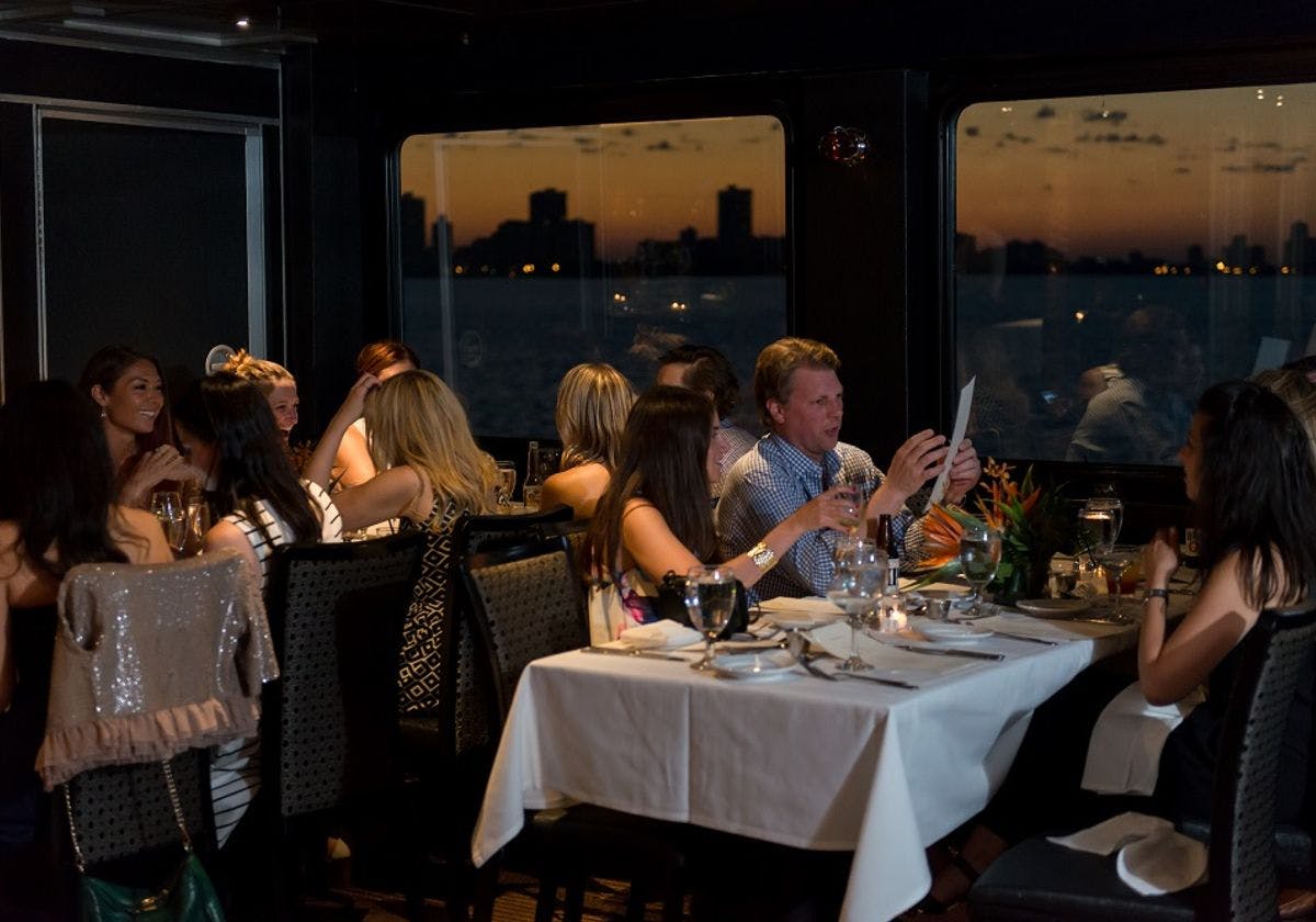 People dining at elegantly set tables in a dimly lit restaurant with large windows and a city skyline at sunset in the background.