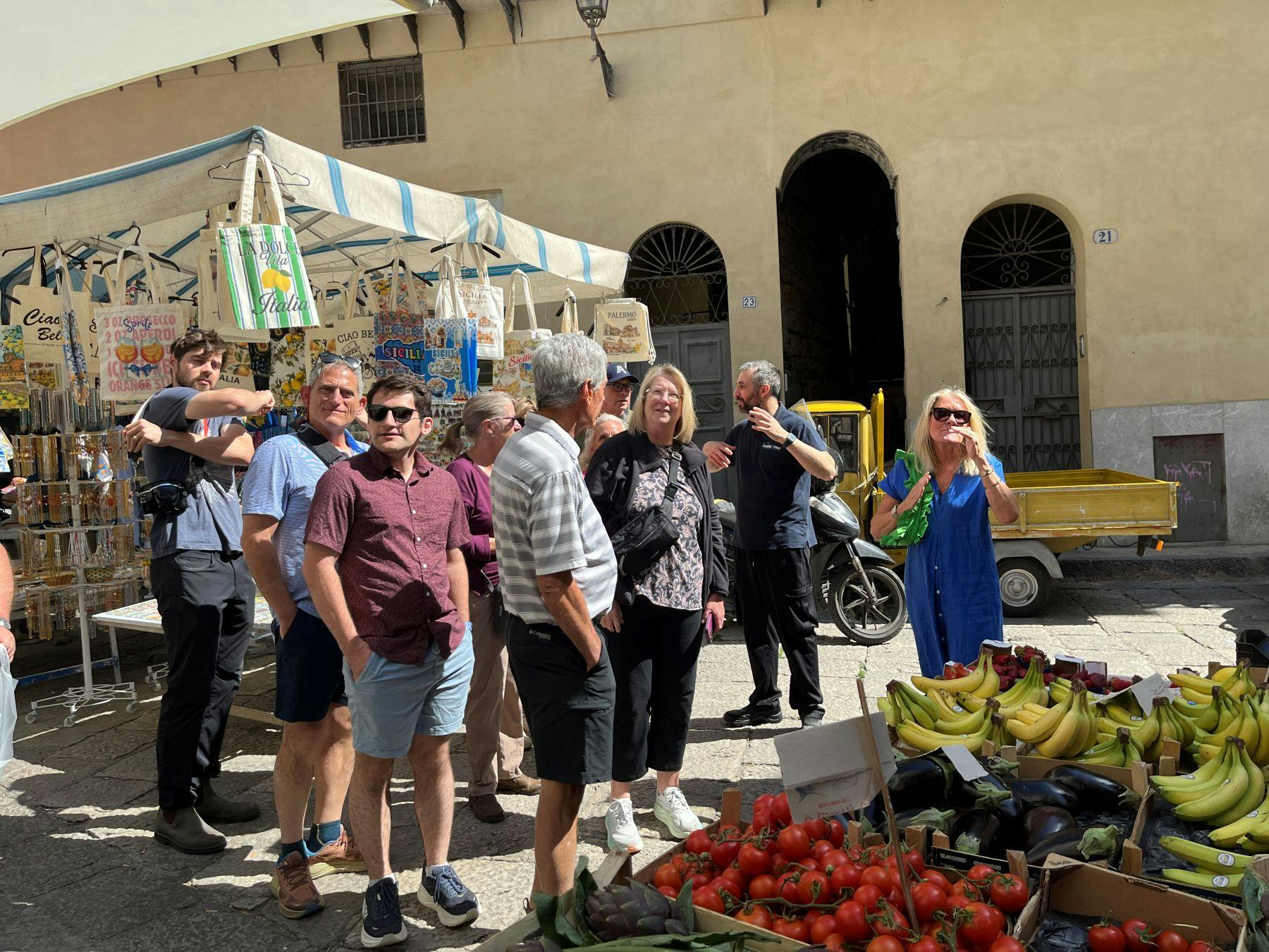A group of people standing near a market stall with colorful items and produce in an outdoor setting.