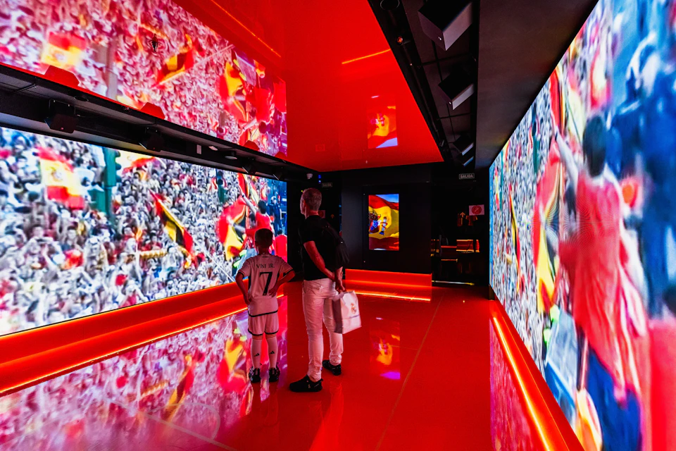 Two people stand in a brightly lit, red room with large screens showing sports scenes and crowd images.