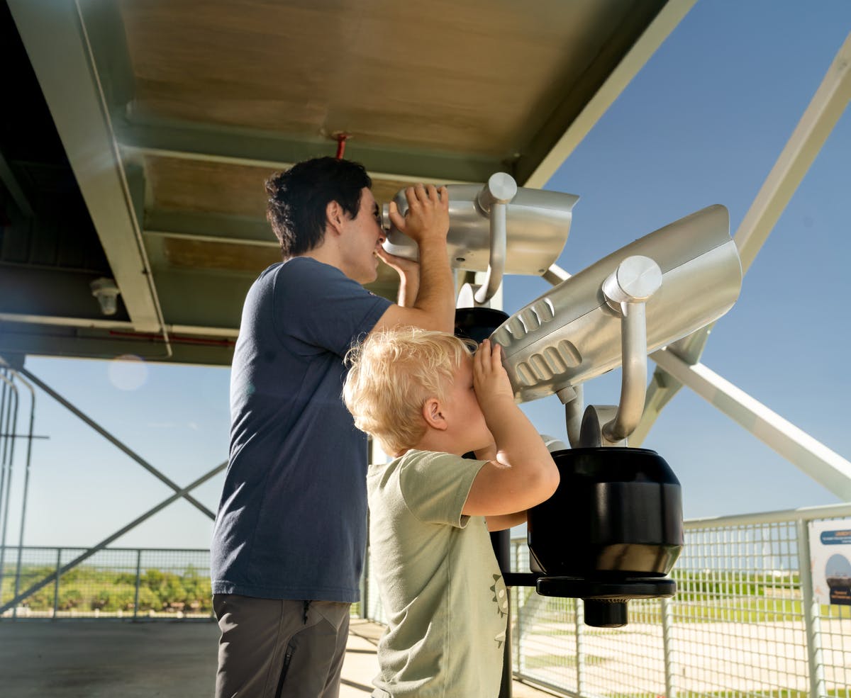 An adult and a child look through coin-operated binoculars on an observation deck with a view of a landscape in the background.