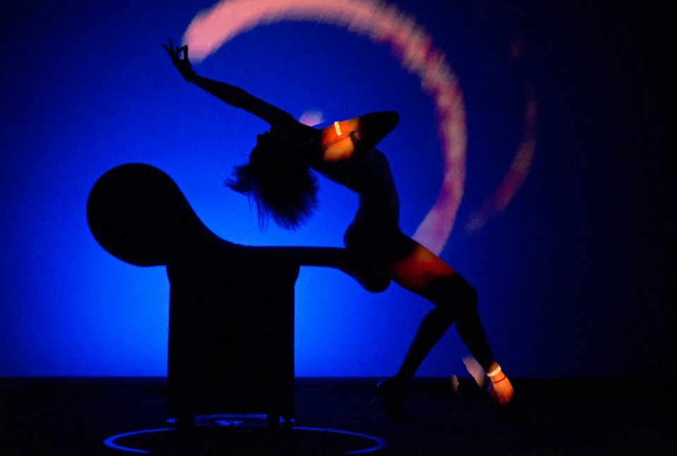 A person striking an expressive dance pose in silhouette against a vibrant blue and magenta background, with a chair.