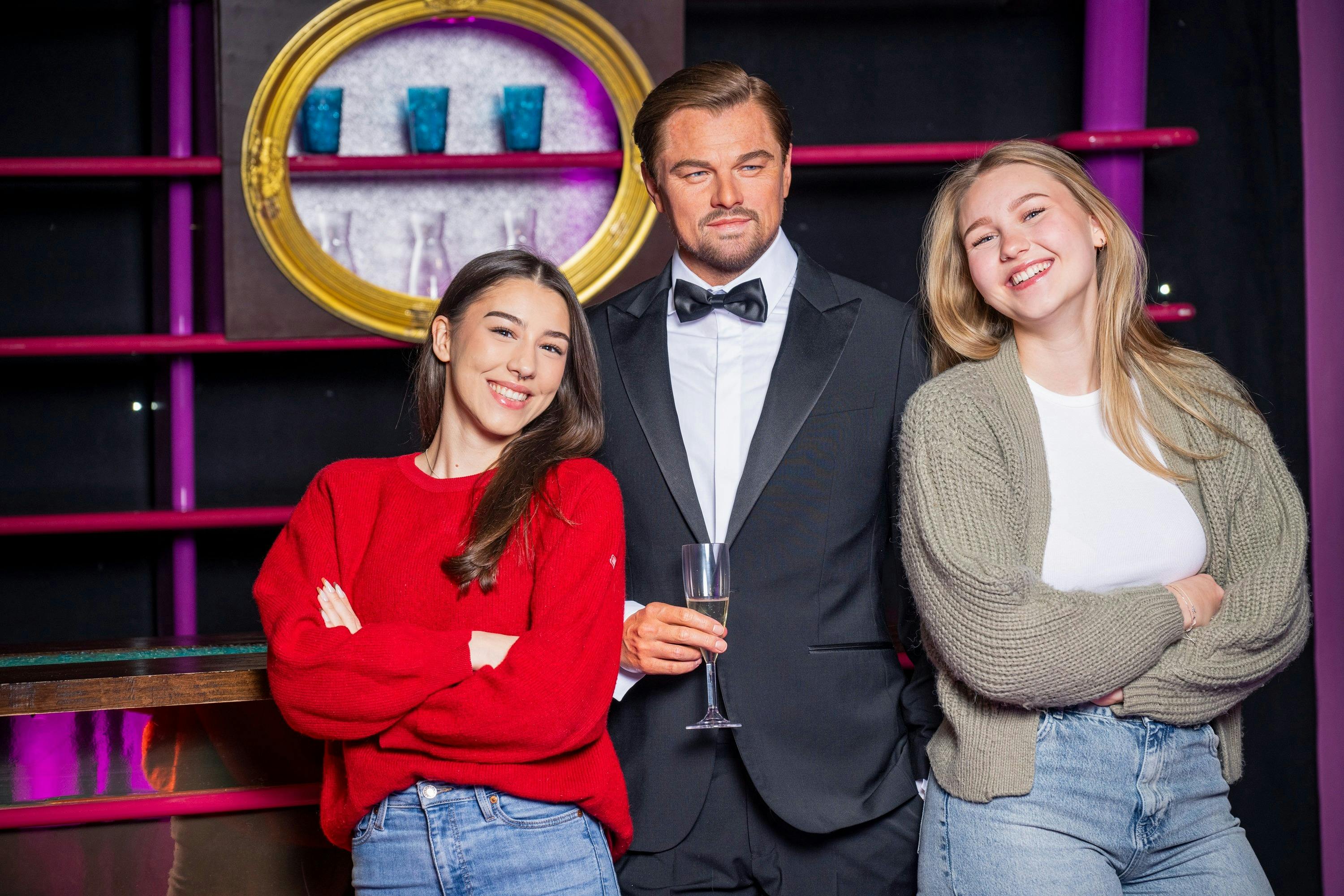 To unge kvinder poserer smilende ved siden af en voksfigur i en smoking, der holder et champagneglas, med en dekorativ baggrund.