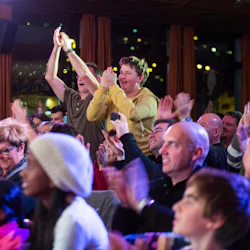 En mångsidig publik som klappar och jublar vid ett inomhusevenemang, med två personer som står och applåderar entusiastiskt.