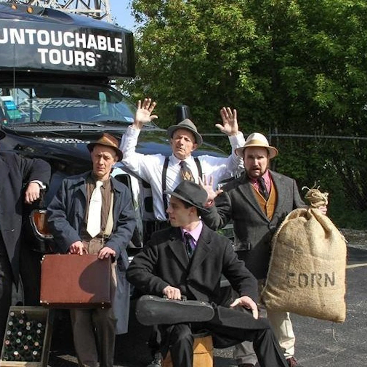 Five people in vintage attire posing with a sack labeled "Corn" and a suitcase in front of a vehicle with "Untouchable Tours."
