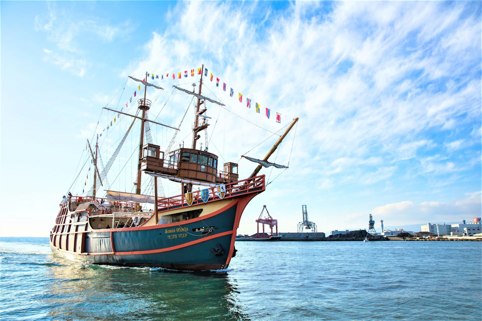 A colorful ship with flags docked on water, with an industrial background and a partly cloudy blue sky.