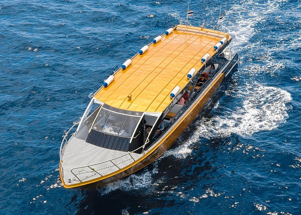 A boat with a yellow roof sails on blue water. Several people are visible inside the boat. Flags are attached to the back.