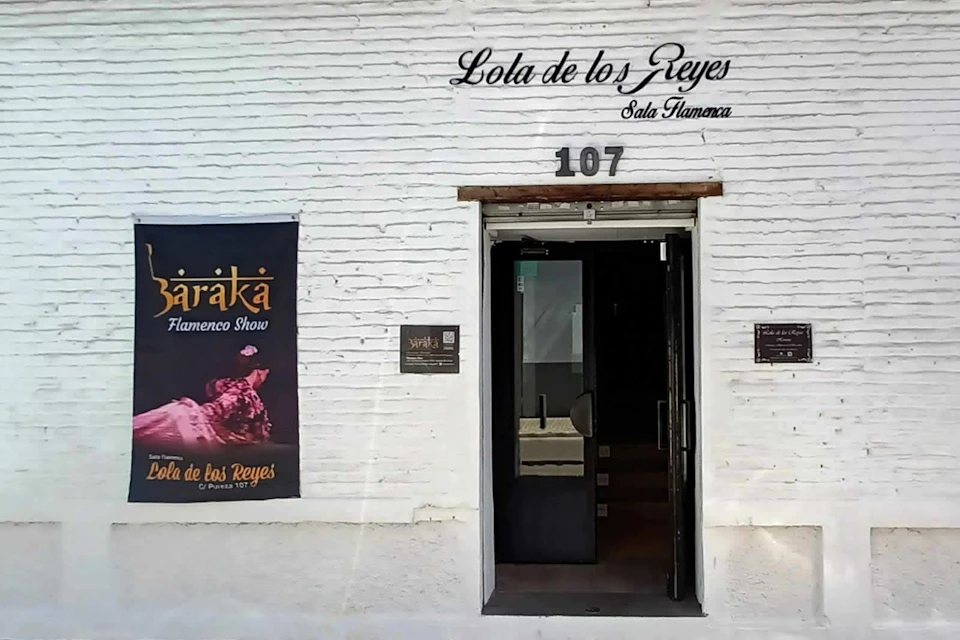 A white brick building entrance with "Lola de los Reyes Sala Flamenca" written above, a poster on the left, and the number 107 above the door.