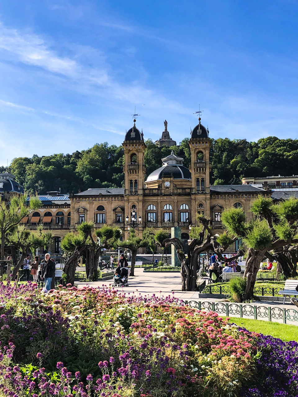 Historic building with twin towers, gardens, and people walking or sitting on benches, under a clear blue sky with a hill in the background.