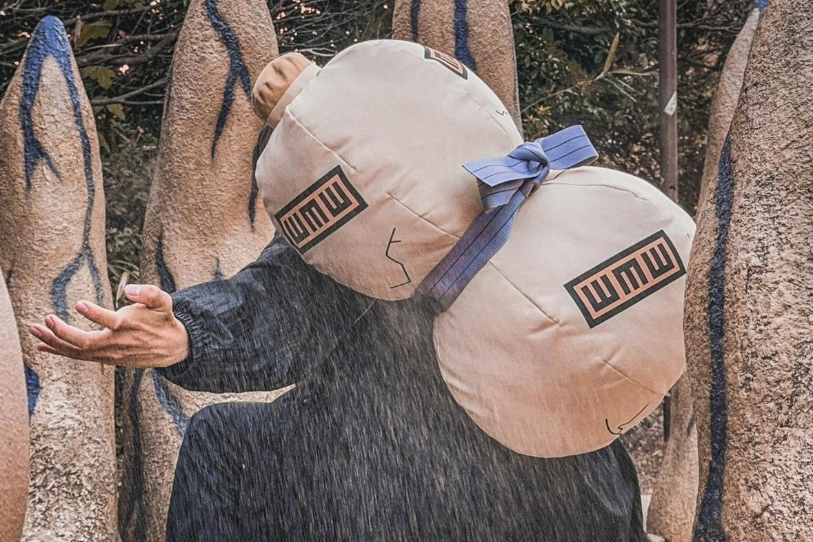 A person is wearing a large sandbag prop on their back with a blue ribbon and markings, in an outdoor setting.