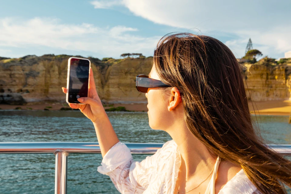 A woman on a boat taking a photo with a smartphone, with cliffs and water visible in the background.
