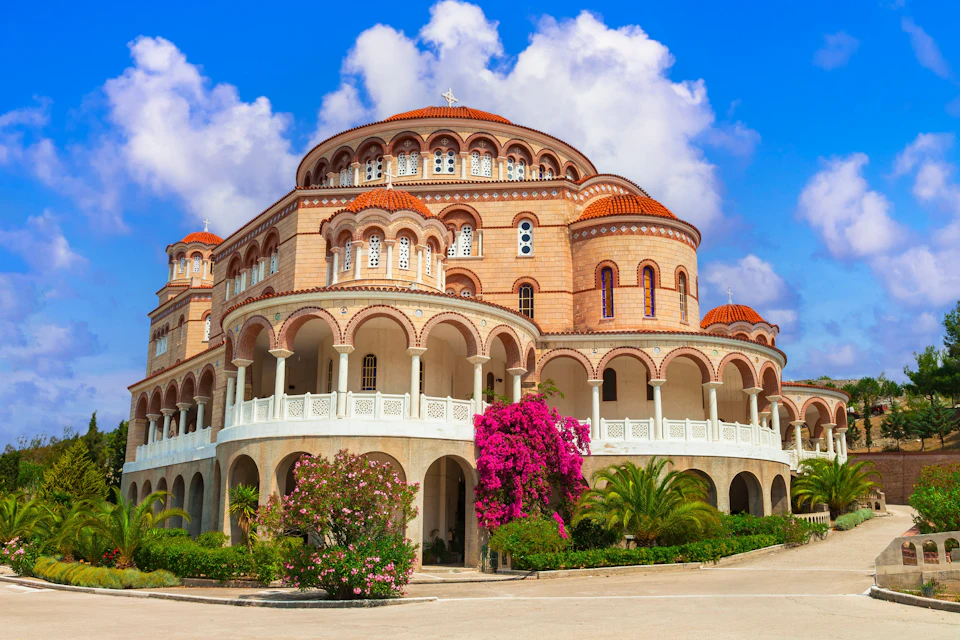 A large ornate building with arches, domes, and a white facade, surrounded by colorful flowers and greenery under a blue sky.
